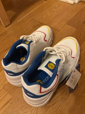 Oanvända Lidl x Esmara vita chunky sneakers  - Säljer ett par vita Lidl x Esmara sneakers med blå, röda och gula detaljer. Skorna har chunky sula men är superlätta och inte tunga att ha på, vita skosnören och blå insida. Märkets logga syns på plösen och innersulan. Tillverkade i syntetmaterial och har en sportig retrostil. Lappar kvar - aldrig använda. Storlek 38.