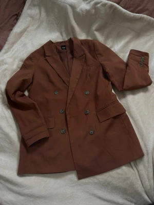 Brun dubbelknäppt kavaj från Sinsay - Snygg brun kavaj från Sinsay i storlek M. Dubbelknäppt modell med fyra knappar framtill och klassisk krage. Kavajen har två fickor med lock och är helfodrad för extra komfort. Perfekt för dig som vill ha en stilren och trendig look.