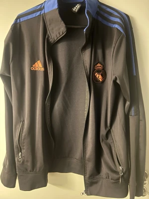 Real Madrid träningsdress Adidas - Snygg svart tränings tracksuit från Adidas med Real Madrid-logga på bröstet och blå detaljer på axlarna.  tracksuit har dragkedja, två fickor med dragkedja och klassiska Adidas-ränder. Perfekt för fotbollsträning eller chill.