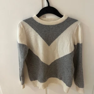 Grå och vit stickad tröja med mönster - Superskön stickad tröja i grått och vitt med V-format mönster framtill. Rund hals och långa ärmar. Perfekt att slänga på sig när det är lite kyligt ute. Saknar lapp så vet tyvärr ej vart den är ifrån, köpte den från Plick men passade tyvärr ej.