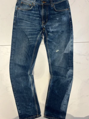 Blå raka jeans från Nudie Jeans - Säljer ett par klassiska blå jeans från Nudie Jeans med sjuk wash rak passform och snygg slitning. Jeansen har fem fickor, kopparfärgade nitar och kontrastsömmar. Tillverkade i kraftig denim med en cool tvättad look och patch bak i midjan.