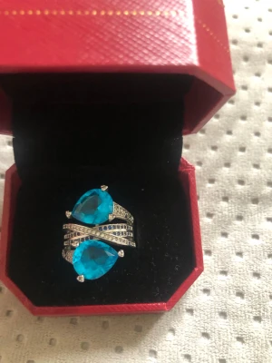 Silverring med blå stenar - Unik silverfärgad ring med två stora, droppformade blå stenar och gnistrande små diamanter på bandet. Ringen har en modern, korsad design och är prefekt för dig som vill sticka ut med ett färgstarkt smycke. Nypris var 2500 kr. 