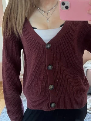 Vinröd kofta från Pieces - Mysig vinröd kofta från Pieces med grovstickad struktur och fyra stora knappar framtill. Koftan har v-ringning och långa ärmar, perfekt att slänga över en topp. Passar dig som gillar en avslappnad men ändå stilren look.