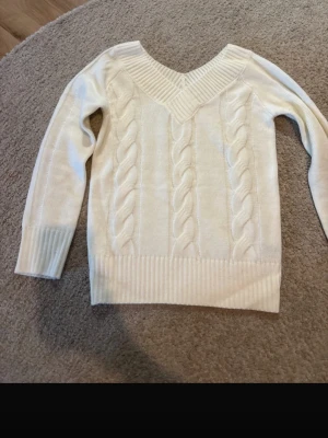 Vit kabelstickad tröja med V-ringning - Säljer en krämvit kabelstickad tröja med bred V-ringning och ribbade muddar. Tröjan har ett klassiskt flätmönster framtill och långa ärmar. Perfekt för lager på lager och en clean look.