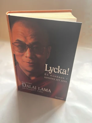 Lycka! Dalai Lama handbok - En inspirerande bok med titeln 'Lycka! En handbok i konsten att leva' av Dalai Lama och Howard C. Cutler. Boken har hårda pärmar i svart och orange med tydlig text på omslaget. Perfekt för dig som vill fördjupa dig i livsfilosofi och personlig utveckling.