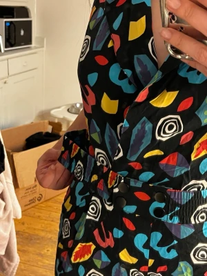 Färgglad kort jumpsuit med mönster - Säljer en svart jumpsuit med färgglada abstrakta mönster i blått, rött, gult och vitt. Från stranger things. Jumpsuiten har korta ärmar, V-ringning och shorts, samt ett elastiskt midjeband med knappar. Perfekt för dig som gillar att sticka ut med din stil!