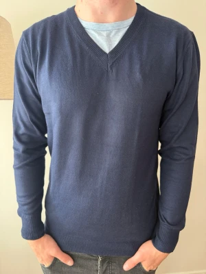 Mörkblå stickad tröja  - En stilren mörkblå stickad tröja med V-ringad hals och långa ärmar. Tröjan har ribbade muddar vid ärmslut och nederkant. Perfekt att bära över en t-shirt för en avslappnad look. Materialet känns mjukt och lätt.