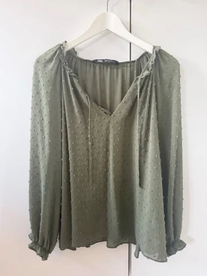 Grön prickig blus från Zara - Luftig grön blus från Zara med diskret prickigt strukturmönster. Blusen har v-ringning med knytband och långa, lösa ärmar med resår vid ärmsluten. Tillverkad i ett tunt, lätt material som ger ett avslappnat intryck.💓 superfint olivgröb färg. Annvänd väldigt lite. Storlek M men passar även en S 