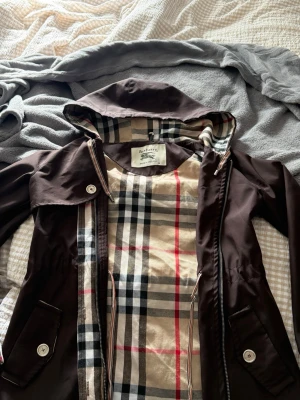 Brun regnjacka från Burberry med huva - Snygg brun regnjacka från Burberry med klassiskt rutigt foder i beige, svart, rött och vitt. Jackan har huva, dragkedja, knappar och två fickor framtill. Perfekt för regniga dagar och ger en stilren look.