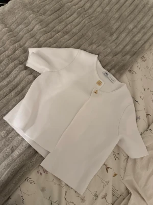 Vit kortärmad kofta från Zara - Stilren vit kofta från Zara med korta ärmar och rund halsringning. Koftan har en rak passform och stängs med två dekorativa guldfärgade knappar framtill. Materialet är mjukt och lätt ribbat, perfekt för en clean och modern look. Storlek S aldrig använd.