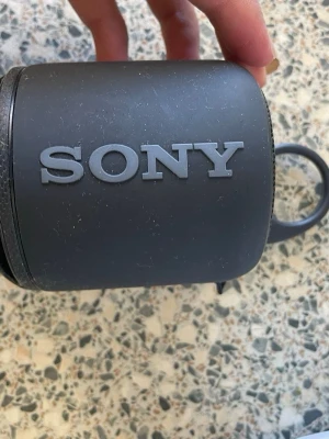 Sony bärbar högtalare svart - Smidig och kompakt bärbar högtalare från Sony i svart färg. Rund form med tydlig Sony-logga och praktisk rem för enkel transport. Tillverkad i tålig plast, perfekt för att ta med sig ljudet överallt.