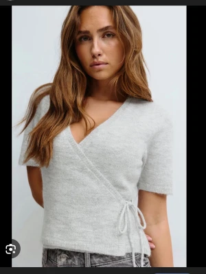 Ljusgrå omlottopp med knytband - Supersöt ljusgrå omlottopp i mjukt, fluffigt material. Knytband i sidan och korta ärmar ger en avslappnad men ändå chic look. Perfekt till jeans eller kjol för en trendig vibe. Djup v-ringning framtill och enkel, stilren design.