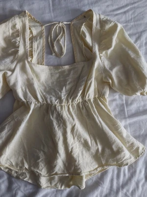 Ljusgul blus med puffärm och knyt - Står s men passar xs-s. Supersöt ljusgul blus i luftig bomull med puffärmar, fyrkantig ringning och knyt i ryggen. Blusen har en lätt volang nedtill och öppen rygg med dekorativ rosett. Perfekt för dig som gillar romantisk och trendig stil.
