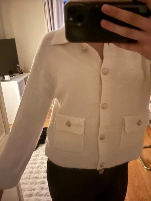 Vit kofta med fickor och knappar - Säljer en stilren vit kofta med stora knappar framtill och två praktiska fickor på magen. Koftan har en klassisk krage och är gjord i ett mjukt, stickat material. Perfekt att slänga på sig för en clean och enkel look.