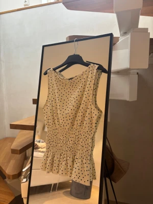 Prickig ärmlös topp med volang - Söt ärmlös topp i benvit med svarta prickar och smockad midja som ger en snygg passform. Toppen har volangkant nedtill och rund halsringning. 