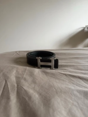 Svart skärp med H-spänne - Snyggt svart skärp med klassiskt H-format metallspänne i silver. Bältet är gjort i slätt skinn och har vita sömmar längs kanterna för en stilren kontrast. Perfekt accessoar för att lyfta vilken outfit som helst.