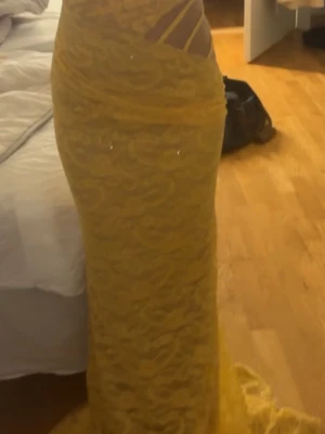 Gul långklänning med spetsdetaljer - Snygg gul långklänning med spetsdetaljer och transparenta partier i midjan. Klänningen har en figurnära passform och elegant släp. Perfekt för dig som vill sticka ut med en unik look.