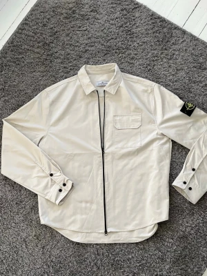 Vit overshirt från Stone Island - Snygg vit overshirt från Stone Island med dragkedja framtill, bröstficka och klassisk avtagbar logotyp-patch på ärmen. Overshirten har knappade manschetter och är tillverkad i ett slätt bomullsmaterial. Perfekt för en clean och stilren look.