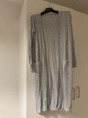 Ljusgrå lång kofta från Lager 157 - Lång och öppen ljusgrå kofta från lager 157 med två fickor framtill. Koftan har långa ärmar och är tillverkad i ett mjukt, tunt material som passar perfekt för lager på lager. Enkel och stilren design som funkar till det mesta.nästan NY 