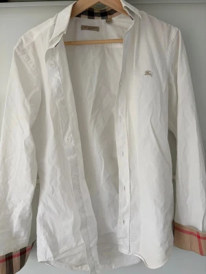 Vit skjorta från Burberry med rutiga detaljer - Klassisk vit skjorta från Burberry med ikoniska beige-rutiga detaljer på insidan av manschetterna och diskret logga broderad på bröstet. Skjortan har lång ärm, normal passform och är tillverkad i mjuk bomull. Perfekt för en clean och stilren look.