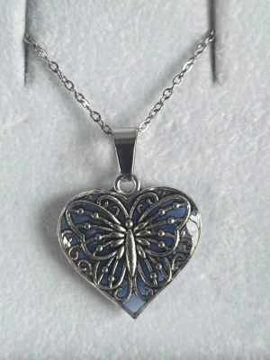 Halsband med hjärthänge och fjäril - Unikt halsband med ett hjärtformat hänge i silverfärgad metall och blå bakgrund. Hänget har ett detaljerat fjärilsmotiv. Kedjan är tunn i rostfritt stål och matchar hängets färg. Perfekt för dig som gillar magiska detaljer och snygga accessoarer. Handgjort. Både kedjan och hänget tål vatten utan att missfärgas.