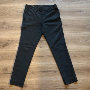 Svarta kostymbyxor från Jack & Jones - Snygga svarta kostymbyxor från Jack & Jones med diskret rutmönster. Byxorna har en klassisk passform med knappar och dragkedja framtill. Perfekta för en stilren look. OBS: Finns två extra knappar till byxan. 