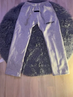 Grå Essentials mjukisbyxor - Säljer ett par grå mjukisbyxor från Essentials med tryckt logga på vänster lår. Byxorna har resår i midjan med dragsko och raka ben. Perfekta för chill dagar och riktigt skön passform. Tillverkade i mjukt material som känns bekvämt hela dagen.