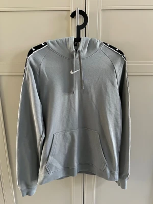 Grå Nike hoodie med logga på ärmar - Snygg grå hoodie från Nike med huva och dragsnören. Klassisk vit Nike-logga på bröstet och svarta band med vita swoosh-loggor längs ärmarna. Stor känguruficka framtill och ribbade muddar. Perfekt för en sportig och avslappnad stil.