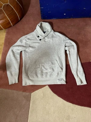 Grå tröja  från Ralph Lauren - Snygg grå sweatshirt från Ralph Lauren med hög krage och två knappar vid halsen. Tröjan har en stor magficka och en patch med PRL & CO-logga nedtill. Tillverkad i mjukt sweatshirtmaterial, perfekt för chill dagar. Ställ frågor om du undrar något!✅✅