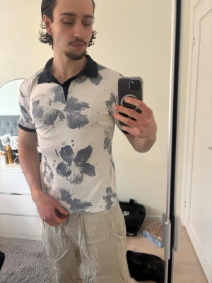 Vit blommig pikétröja med svart krage - Vit pikétröja med stora gråblå blommor och svart krage. Tröjan har korta ärmar med svarta muddar och en klassisk pikéknäppning. Perfekt för en avslappnad men stilren look.