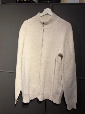 Beige stickad ziptröja med hög krage - Säljer en stilren beige stickad ziptröja med hög krage och långa ärmar. Tröjan har en diskret struktur i materialet och dragkedja framtill. Perfekt för lager på lager och enkel att matcha med olika outfits.