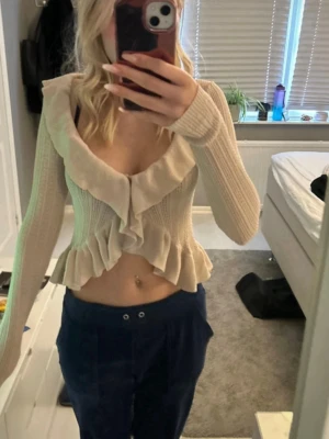Beige volangstickad kofta - Säljer en beige stickad kofta med volangdetaljer längs urringningen och nederkanten. Koftan är croppad, har långa ärmar och är tillverkad i ett mjukt, ribbat material. Perfekt för dig som gillar en söt och trendig stil.