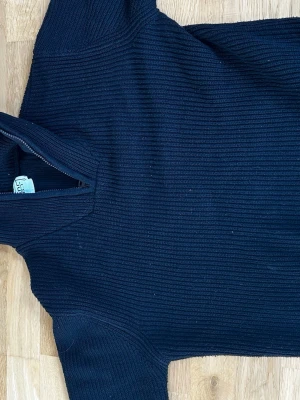 Mörkblå stickad halvzip tröja - Mörkblå stickad tröja med halvzip och hög krage. Tröjan har ribbad struktur och långa ärmar, perfekt för kyliga dagar. Klassisk och enkel design som passar till det mesta.