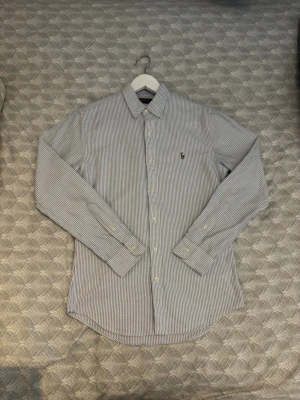 Randig skjorta från Ralph Lauren - Klassisk blåvit randig skjorta från Ralph Lauren med broderad logga på bröstet. Skjortan har lång ärm, knappar framtill och krage. Tillverkad i bomull för en skön och stilren känsla. Den är inte alls använd mycket och är i utmärkt skick!