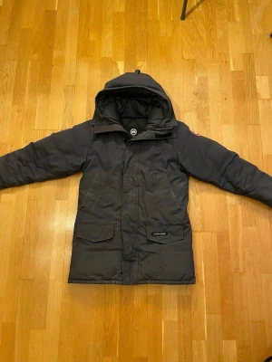 Svart parkas från Canada Goose - Säljer en svart parkas från Canada Goose med klassisk patch på ärmen och stor huva. Jackan har två stora fickor framtill och dragkedja med vindslå. Perfekt för kalla dagar och riktigt snygg streetstil.