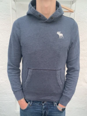Blå hoodie från Abercrombie & Fitch - Snygg blå hoodie från Abercrombie & Fitch med klassisk känguruficka och huva. På bröstet finns en vit broderad älglogga. Hoodien har ribbade muddar vid ärmslut och nederkant. Perfekt för en avslappnad och stilren look.