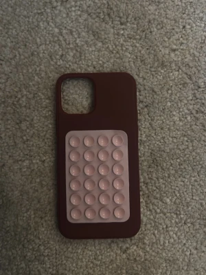 Brunt mobilskal med sugpropp-detalj iPhone 12 - Säljer ett vinrött mobilskal med en ljusrosa sugpropp-panel på baksidan. Skalet är gjort i mjukt silikonmaterial och har en rektangulär form med stora kamerauttag. Perfekt för dig som gillar att pilla på mobilen och vill ha något unikt.