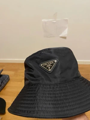 Svart bucket hat  Prada - Stilren svart bucket hat från Prada med klassisk trekantig logotyp framtill. Hatten är tillverkad i ett glansigt, vattenavvisande material som ger en lyxig känsla. Perfekt accessoar för att lyfta din streetwear-look.Likande modell som orginal 