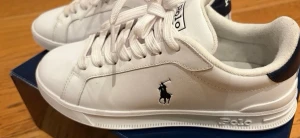 Vita Polo Ralph Lauren sneakers - Stilrena vita sneakers från Polo Ralph Lauren med klassisk låg profil. Skorna har vit ovandel i läder, vit sula och detaljer i mörkblått på hälen. Ikoniska Polo-loggan i svart på sidan och Polo-tryck på plösen. Perfekta för en clean och sportig look. Pris kan diskuteras storlek 38