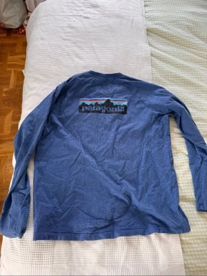 Blå sweatshirt Patagonia tröja - Säljer en blå långärmad tröja från Patagonia med det klassiska logotrycket på ryggen. Tillverkad i en mjuk bomulls- och polyesterblandning som känns skön mot huden. Perfekt för dig som gillar outdoor-vibbar och stilren design. Storlek L och pris kan diskuteras 