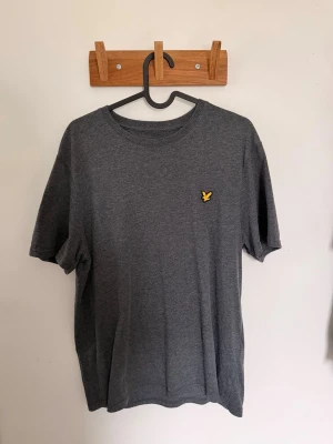 Mörkgrå t-shirt från Lyle & Scott - Mörkgrå t-shirt från Lyle & Scott med klassisk rund hals och korta ärmar. På bröstet finns det ikoniska gula örnloggan broderad. T-shirten är i mjukt bomullsmaterial och har en enkel, stilren design som passar till det mesta.