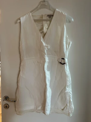 Vit omlottklänning  - Vit ärmlös kortklänning i linne med omlott och v-ringning. Klänningen har en snygg detalj med spänne i sidan och dragkedja bak. Perfekt för sommarens varma dagar och enkel att styla med smycken eller sandaler.
