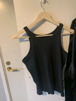 Svart linnetopp i bomull - En stilren svart linnetopp med breda axelband och rundad halsringning. Toppen är tillverkad i mjuk bomull och har en klassisk, tight passform som passar perfekt till jeans eller kjol.