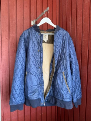 Blå quiltad pufferjacka Timberland - Snygg blå pufferjacka från Timberland i quiltad design med ribbade muddar och krage. Jackan har två fickor med beige detaljer och ett mjukt, ljust innerfoder. Perfekt för kalla dagar och riktigt skön att bära.