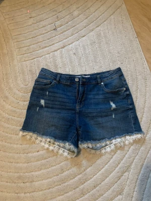 Blå jeansshorts med spets från Amisu - Snygga blå jeansshorts från Amisu med slitna detaljer och vit spetskant längst ner. Klassisk femficksmodell med knapp och dragkedja framtill. Perfekta för en trendig sommarlook.
