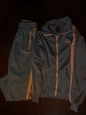 Blått Los Angeles loungewear-set - Snyggt loungewear-set från Bik Bok i ljusblått med vita detaljer och dragkedjor. Setet består av en ziptröja med ståkrage och matchande byxor, båda med 'Los Angeles'-text broderad. Perfekt för chill dagar hemma eller när du vill vara bekväm men ändå se fräsch ut.