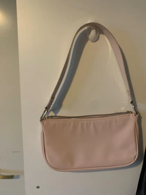 Ljusrosa axelväska från H&M Divided - Söt ljusrosa handväska från H&M Divided med stilren, avlång form och silvriga metalldetaljer. Väskan har ett kort axelband och dragkedja upptill. Tillverkad i slätt syntetmaterial som ger en modern look.