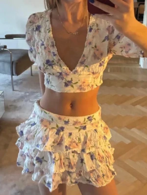 Blommigt set med volanger och puffärm - Säljer ett superfint tvådelat set med blommigt mönster i lila, rosa och grönt. Toppen har djup v-ringning, korta puffärmar och smock i ryggen. Kjolen är kort med flera volanglager och resår i midjan. Perfekt för dig som gillar romantisk och somrig stil.                                                                             KJOL- S, TOPP-XS MEN PASSAR S