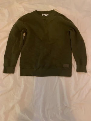 Olivgrön stickad tröja från Zara  - Snygg olivgrön stickad tröja från Zara med diskret patch framtill. Tröjan har rund halsringning, ribbade muddar vid ärmslut och nederkant samt en klassisk rak passform. Perfekt för lager på lager och enkel att matcha.