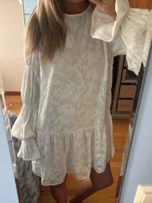 Vit volangklänning  - Superfin vit kortklänning med volangdetaljer och stora vida ärmar. Klänningen har ett subtilt mönster i tyget och en stor rosett framtill. Perfekt till studenten eller sommarn! Vara använd en gång!